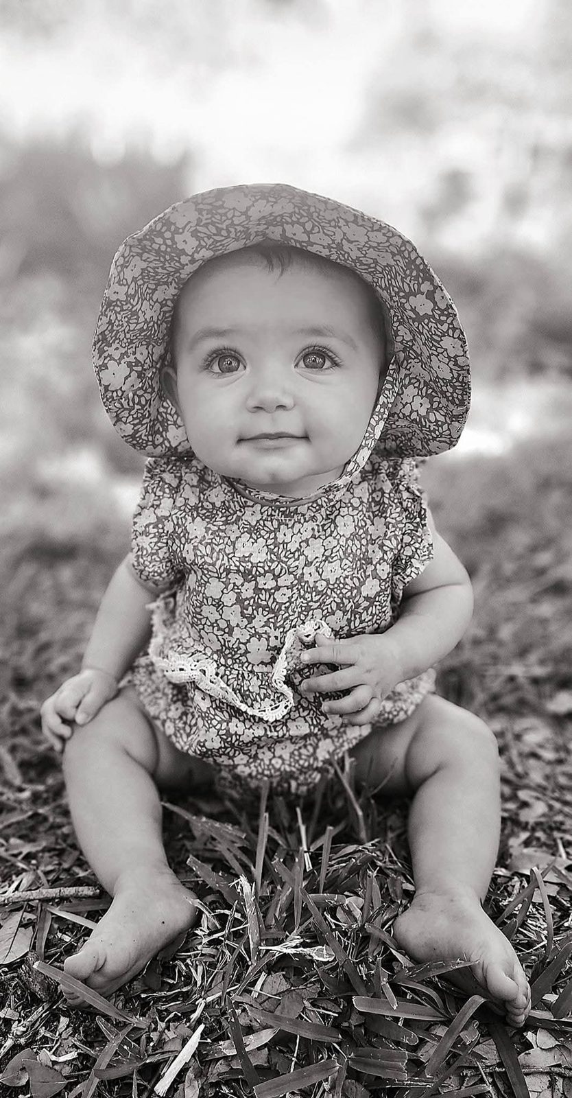 Baby in a floral bonnet sitting in the grass, black and white