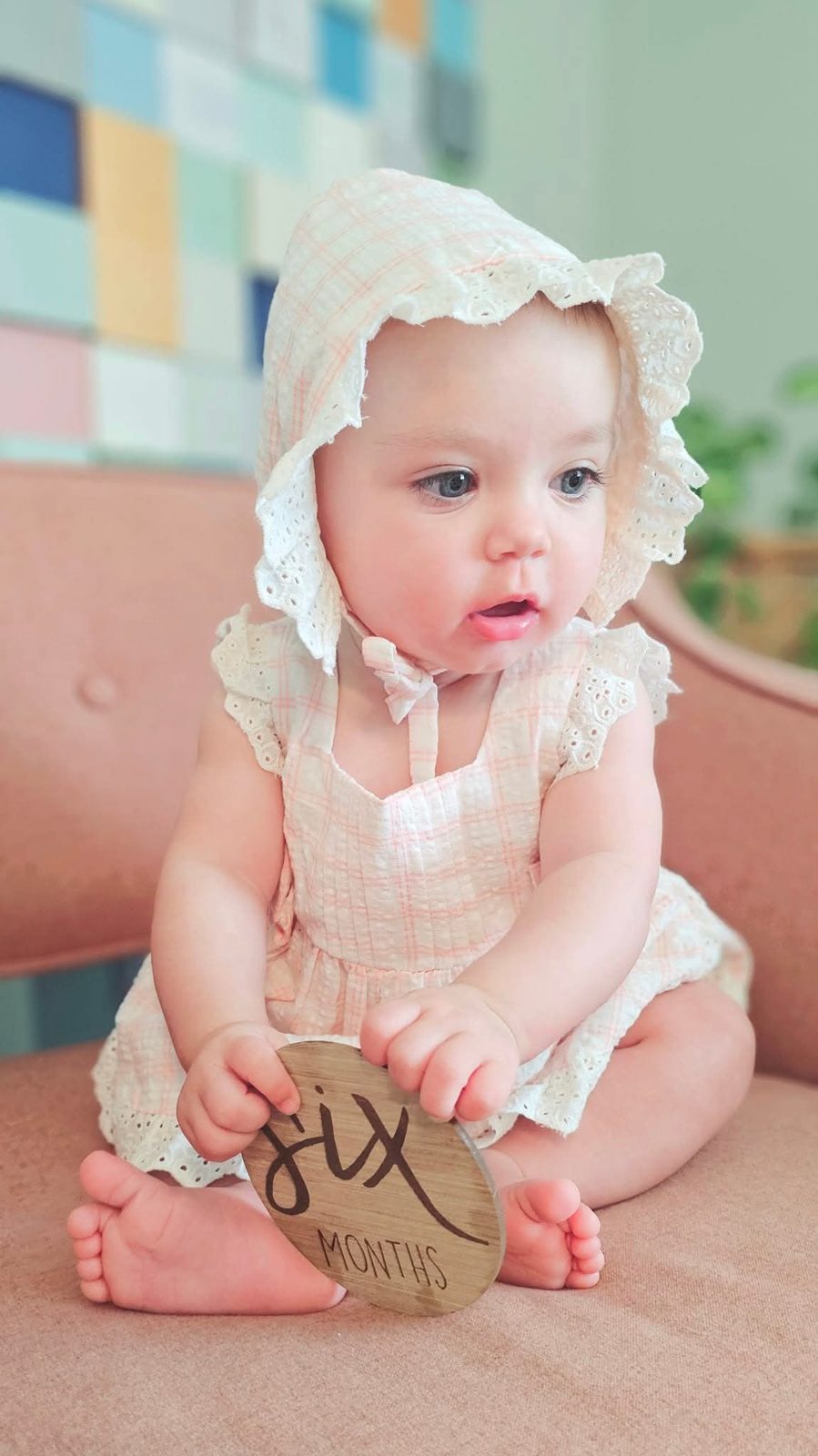 Baby in an eyelet bonnet holding a six months sign