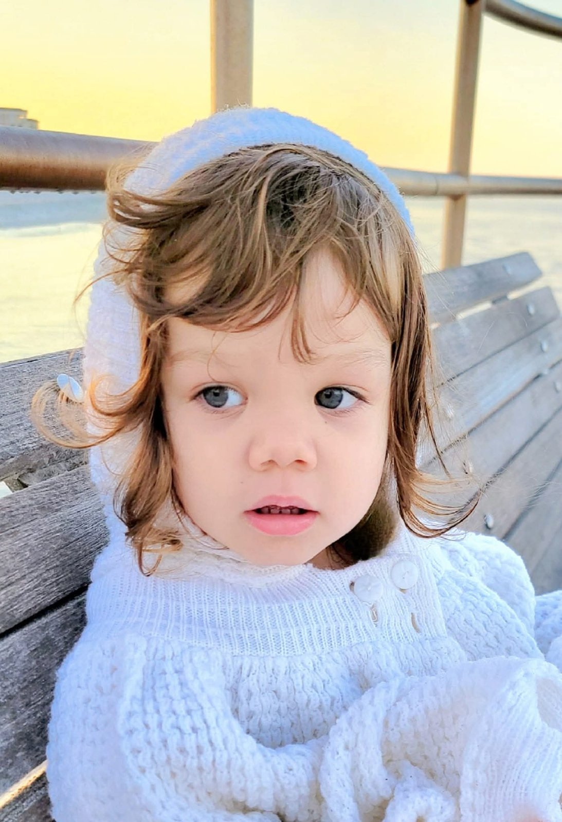 Toddler in a white knit hood on a pier at sunset